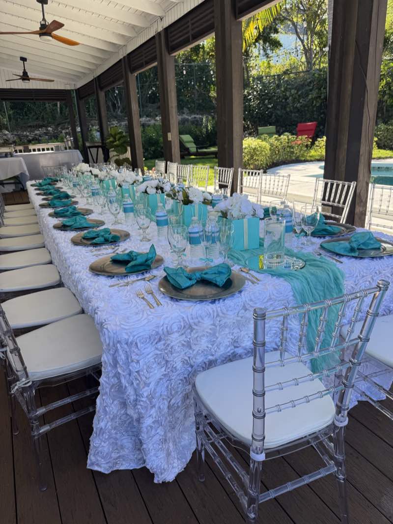 A long table beneath a wooden pergola with teal napkins, white flowers and clear chairs.