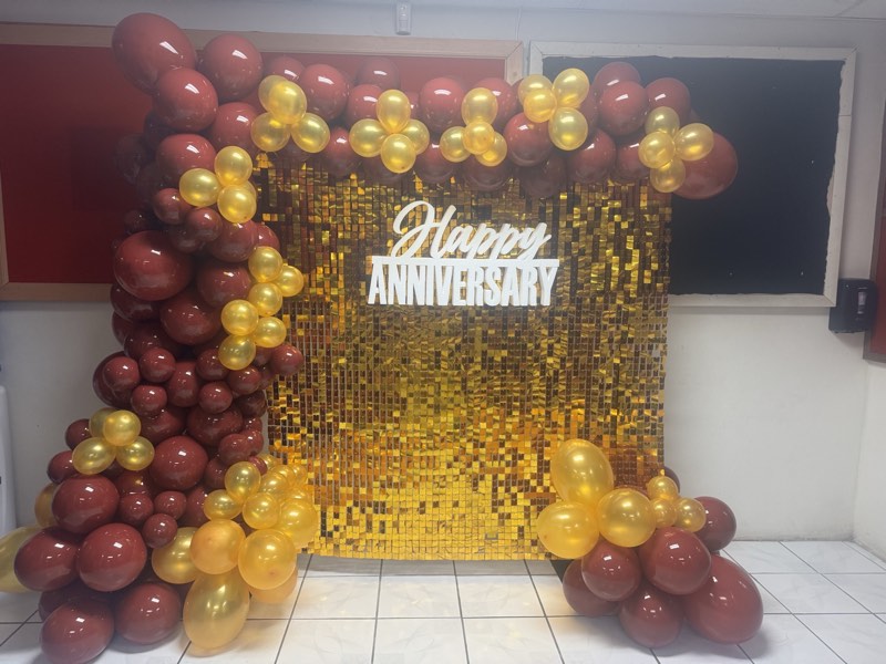 A gold sequin wall with a Happy Anniversary sign framed by burgundy and gold balloons.