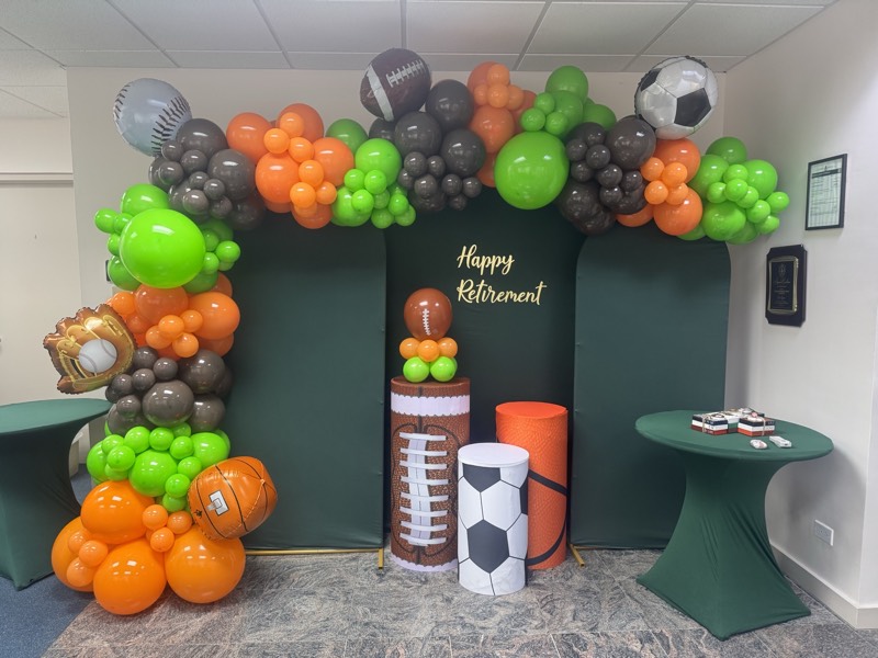 A green Happy Retirement backdrop with orange, green and charcoal balloons and sports-themed plinths.