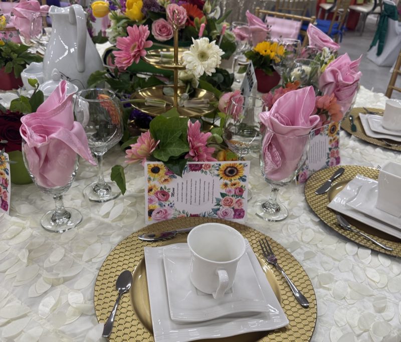 A banquet place setting with a gold charger, white crockery, pink napkins and colourful flowers.