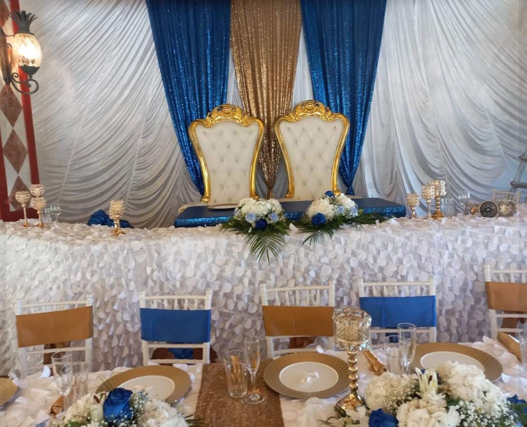 Two gold throne chairs at a white head table in front of blue, gold and white sequin draping.